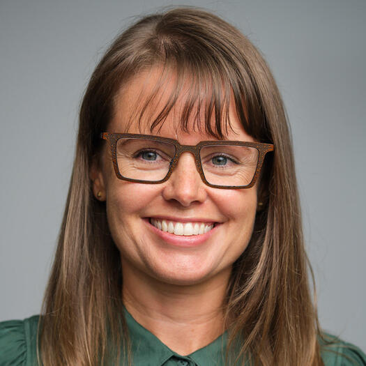 A photo of a person looking to the left, smiling. She is a white person with brown hair, wearing glasses and a green top.